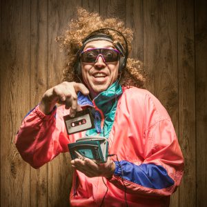 A funky hipster man in late 1980's / early 1990's fashion style, with curly hair and fluorescent colored track suit listening to a walkman tape player and wearing ear muff headphones. He smiles with a cheesy grin, putting a cassette tape into the player with great excitement. Wood paneling in the background. Square crop.