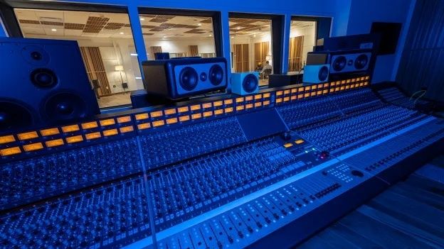 Stor lydmikser i blått lys, omgitt av høyttalere i et studio